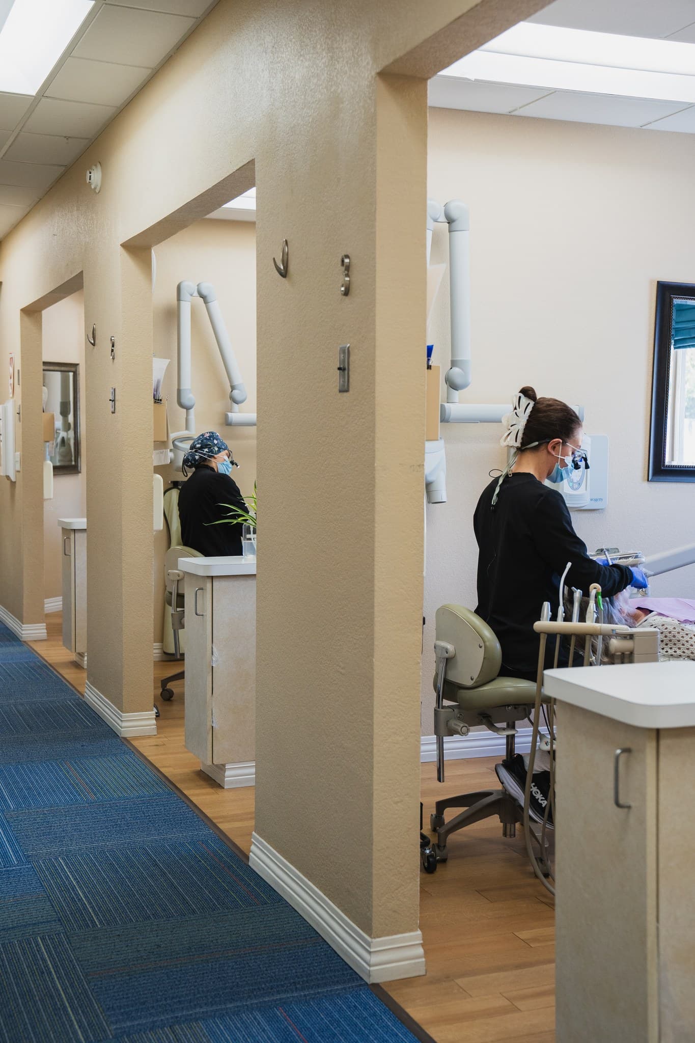 Clinicians in masks and loupes treat patients in open treatment rooms along a hallway at J Street Dental Group, a restorative dentist in Sacramento